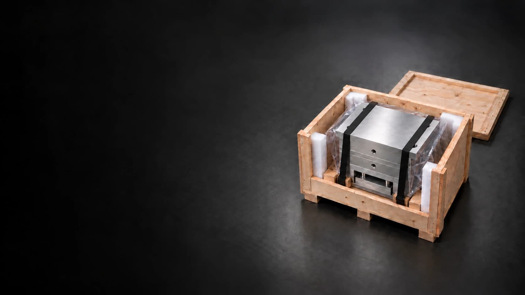 Wooden shipping crate containing the updated mould base, with its corner holes partly covered by black straps, protective wrapping and foam on a dark graphite floor